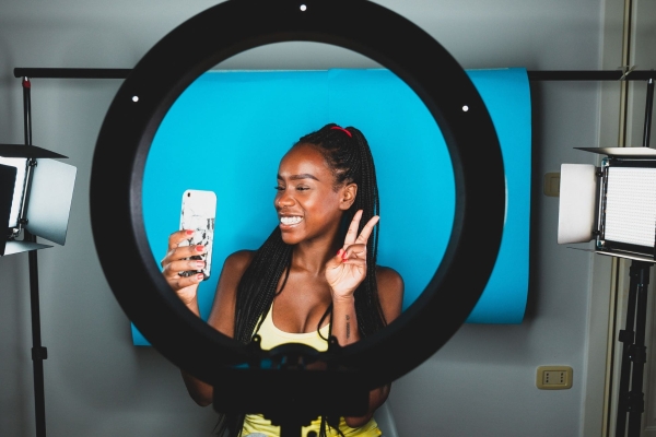 In a studio, a ring light frames a young Black female influencer who is smiling while looking into the mobile phone that she is holding.
